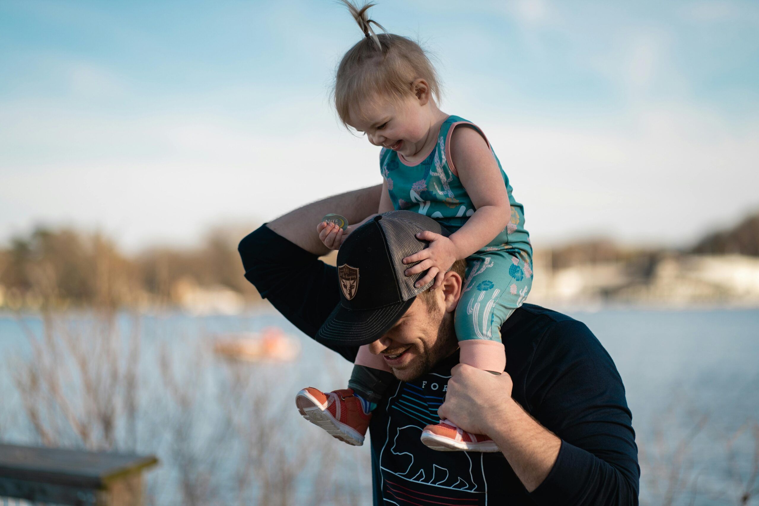 Nurturing Fathers Program A father wearing a black mesh baseball cap supports a baby girl on his shoulders.